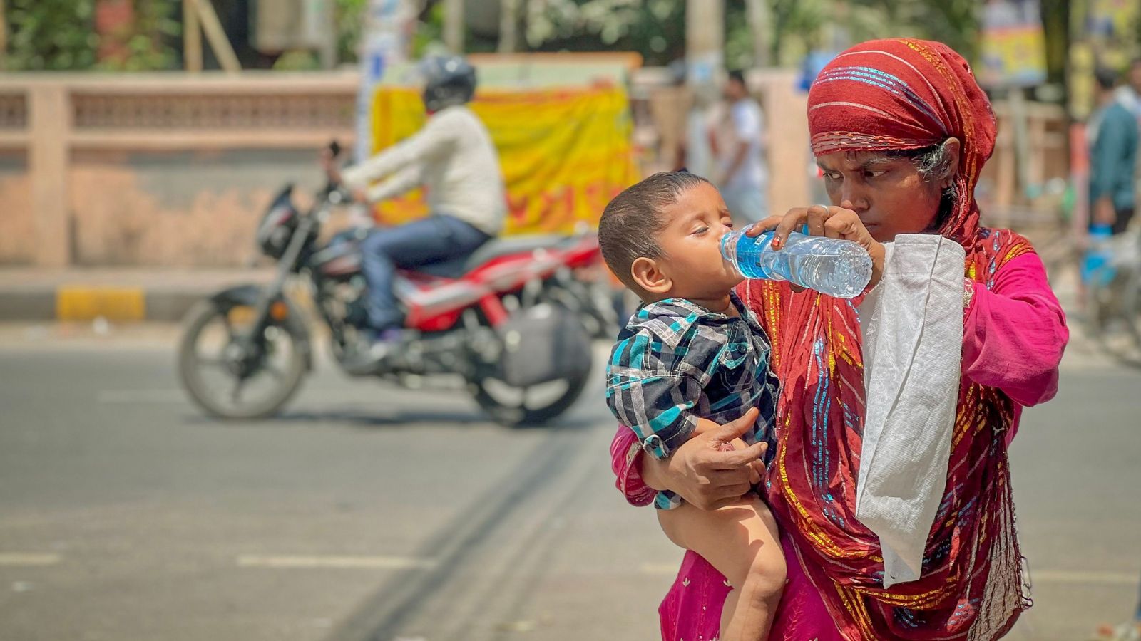 Woman holding a child and offering water during hot weather on a city street, illustrating the human impacts of extreme heat and humidity in India.