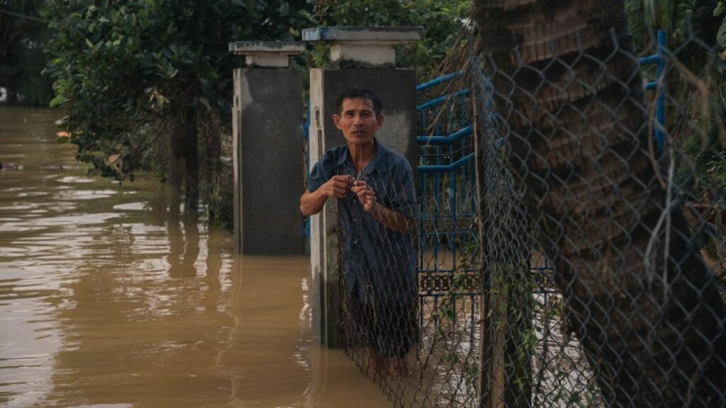 Assam's flood submerges homes as a man stands behind a fence in muddy floodwater.