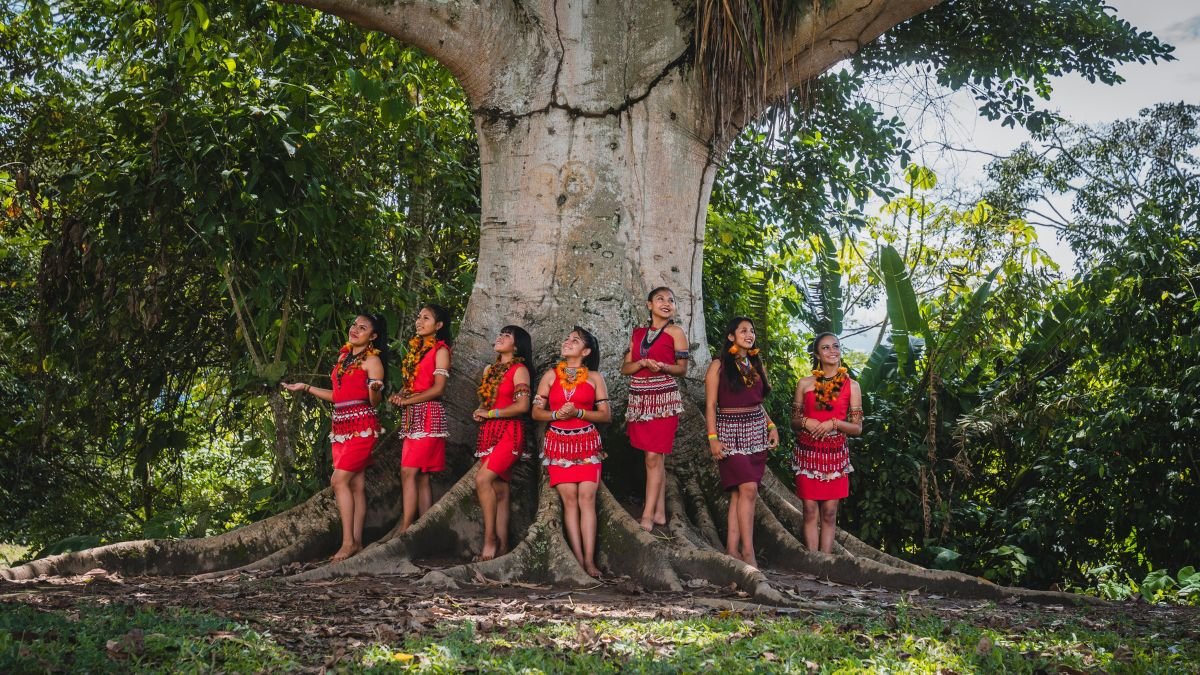 Indigenous women standing beside a large tree in a tropical forest, illustrating the relationship between Indigenous communities and nature conservation