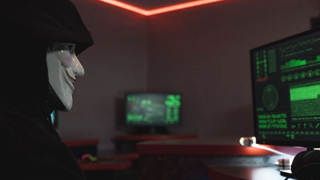 Masked figure facing computer screens, illustrating the growing use of AI tools in cyberattacks such as phishing, stolen-data analysis and cyberattack automation.