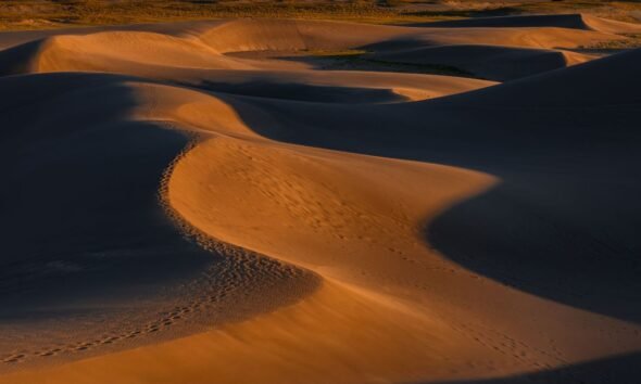 Wind-shaped sand dunes casting long shadows across a desert landscape, with footprints visible along a dune ridge.