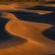 Wind-shaped sand dunes casting long shadows across a desert landscape, with footprints visible along a dune ridge.