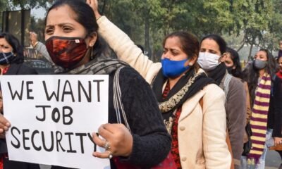 Women wearing masks march in a protest, holding a sign that reads “WE WANT JOB SECURITY.”