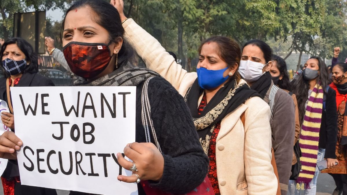 Women wearing masks march in a protest, holding a sign that reads “WE WANT JOB SECURITY.”