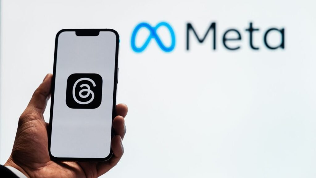 A hand holding a smartphone displaying the Threads app logo, with the Meta logo visible in the background.