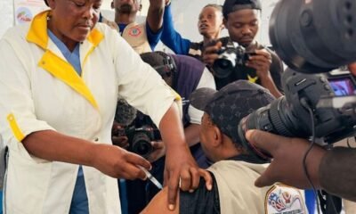 Healthcare worker administers an Ebola vaccine injection to a person as photographers document the vaccination.