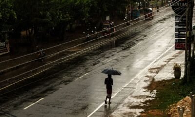 A person walks under an umbrella through light drizzle on a wet Kerala road during a deficient monsoon.