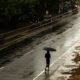 A person walks under an umbrella through light drizzle on a wet Kerala road during a deficient monsoon.