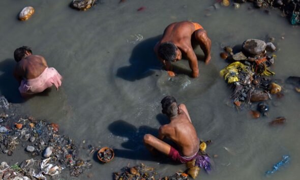 Manual scavenging: Three sanitation workers manually clean a garbage-filled, polluted waterway while standing and crouching in contaminated water, illustrating the hazardous conditions faced by workers involved in manual scavenging and sanitation work.