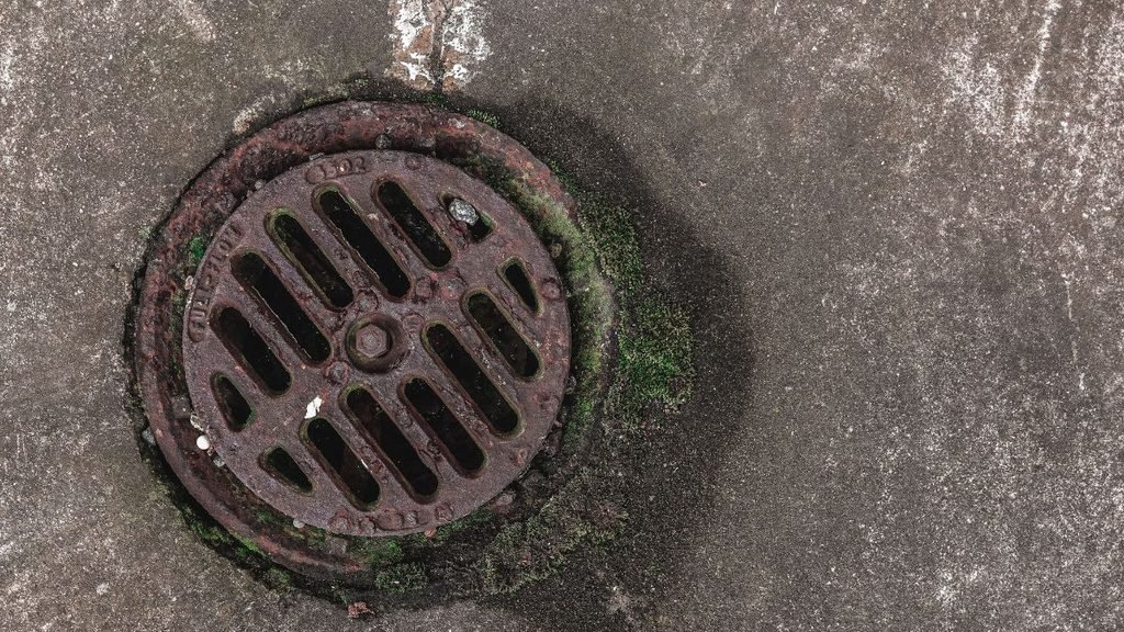 A small, rusted circular manhole cover set into a concrete surface, illustrating the narrow openings through which sanitation workers may be expected to access sewers and confined spaces.