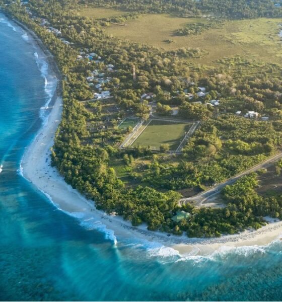 Aerial view of a coastal community in the Marshall Islands, with turquoise waters, sandy beaches, homes, roads and green vegetation along the shoreline.