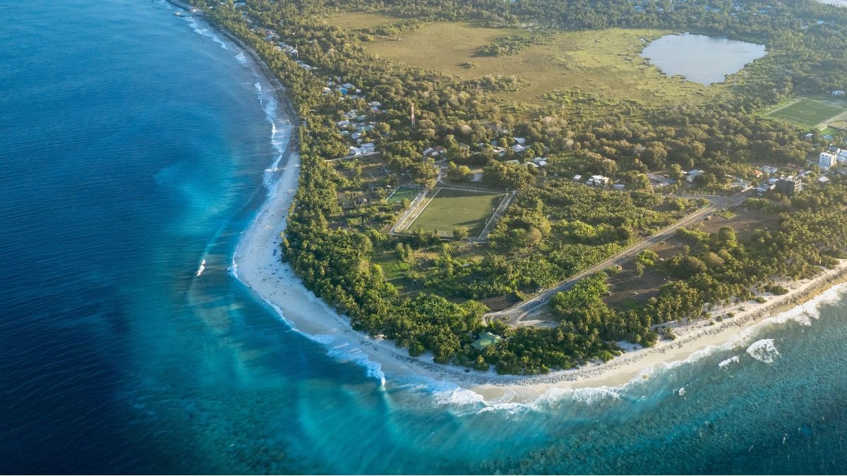 Aerial view of a coastal community in the Marshall Islands, with turquoise waters, sandy beaches, homes, roads and green vegetation along the shoreline.