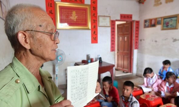Teacher workforce: An older teacher holds a lesson book while teaching a group of children in a classroom.