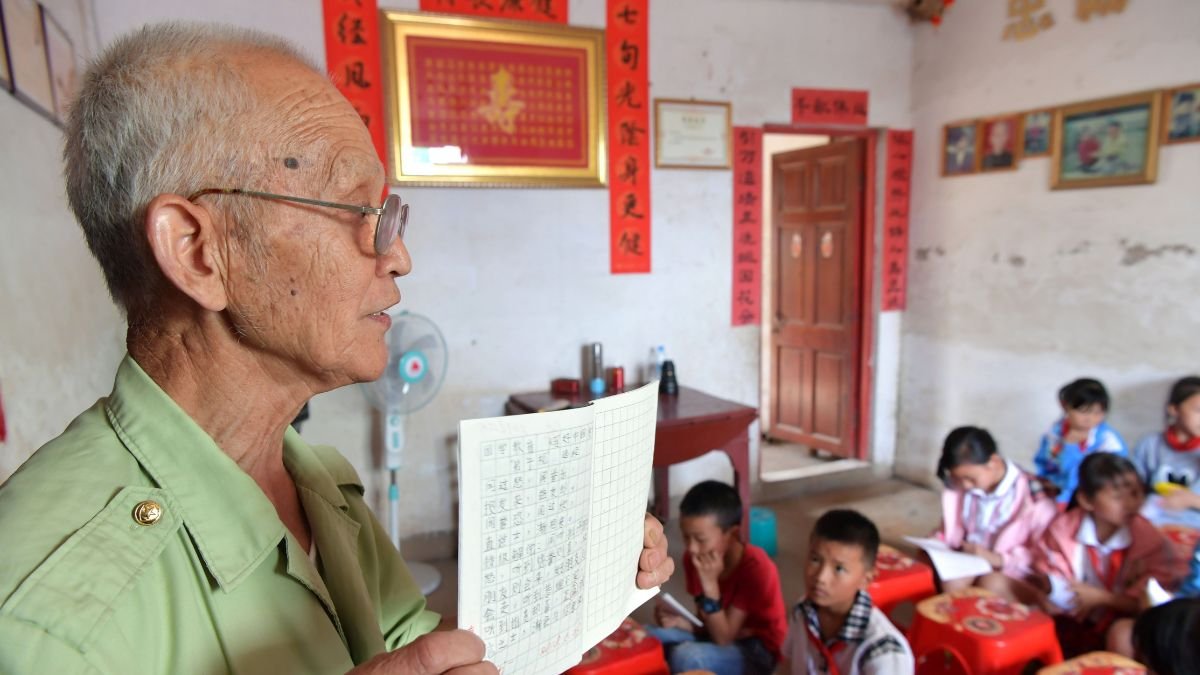 Teacher workforce: An older teacher holds a lesson book while teaching a group of children in a classroom.