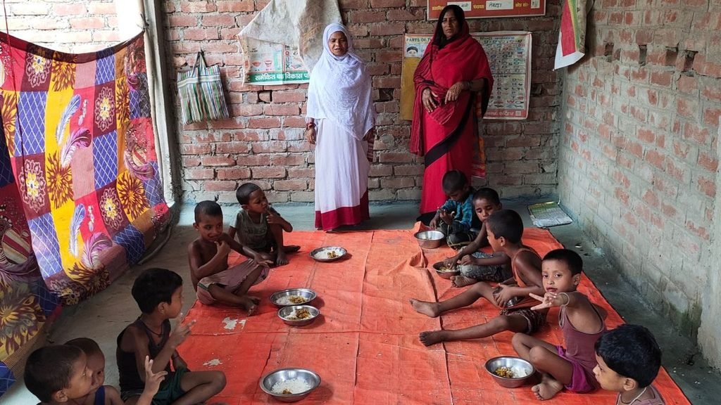 Children eating a school meal while two women stand nearby at an Anganwadi.