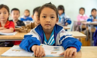 Schoolchildren in a classroom in Northeast India, with a student in a blue-and-white jacket reading a textbook at her desk.