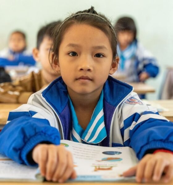 Schoolchildren in a classroom in Northeast India, with a student in a blue-and-white jacket reading a textbook at her desk.