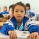 Schoolchildren in a classroom in Northeast India, with a student in a blue-and-white jacket reading a textbook at her desk.