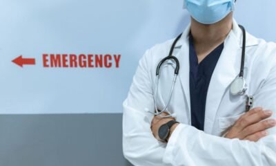 Doctor wearing a surgical mask and stethoscope stands beside an emergency sign in a hospital corridor.