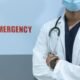 Doctor wearing a surgical mask and stethoscope stands beside an emergency sign in a hospital corridor.