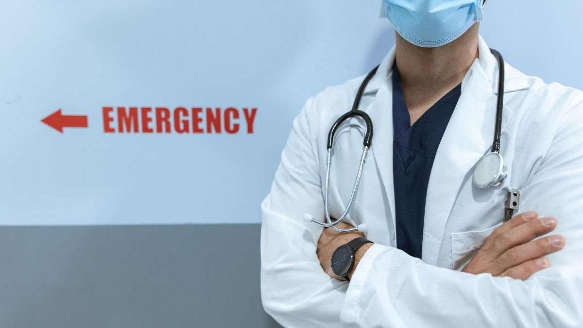 Doctor wearing a surgical mask and stethoscope stands beside an emergency sign in a hospital corridor.