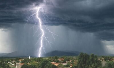 Lightning strikes beneath dark storm clouds as heavy rain falls over a town near green hills.