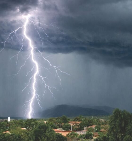 Lightning strikes beneath dark storm clouds as heavy rain falls over a town near green hills.