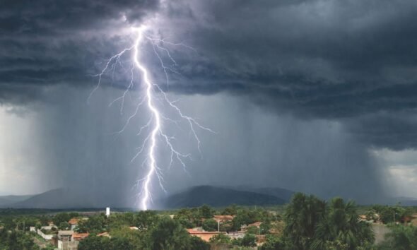 Lightning strikes beneath dark storm clouds as heavy rain falls over a town near green hills.