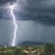 Lightning strikes beneath dark storm clouds as heavy rain falls over a town near green hills.