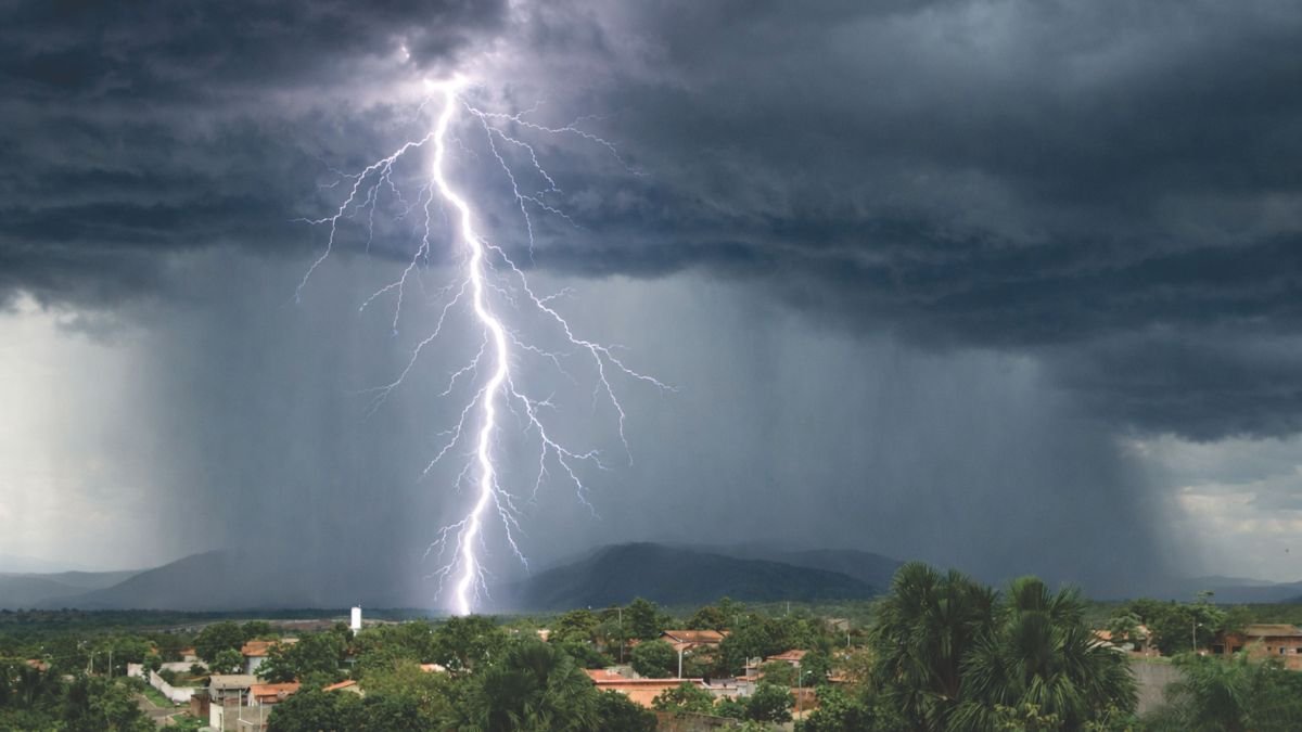 Lightning strikes beneath dark storm clouds as heavy rain falls over a town near green hills.