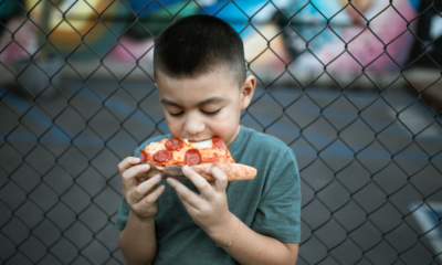 A young boy eating a slice of pepperoni pizza in front of a chain-link fence.