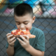 A young boy eating a slice of pepperoni pizza in front of a chain-link fence.