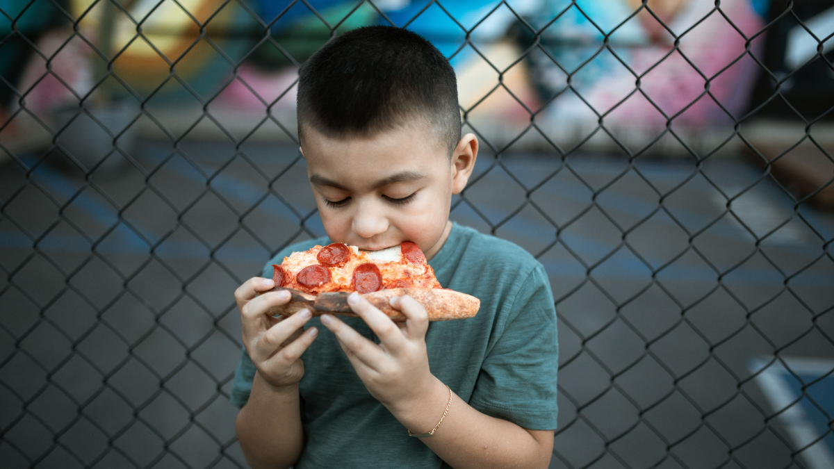 A young boy eating a slice of pepperoni pizza in front of a chain-link fence.