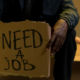 Person holding a cardboard sign reading “Need a Job,” representing unemployment and the global struggle for employment.