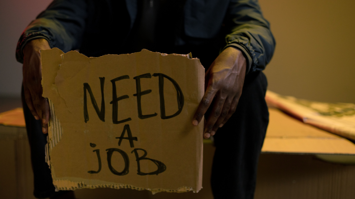 Person holding a cardboard sign reading “Need a Job,” representing unemployment and the global struggle for employment.