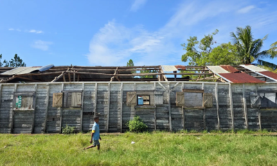 A child walks past a weather-damaged wooden school building with a partially missing roof.