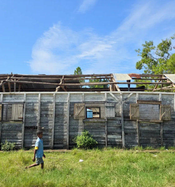 A child walks past a weather-damaged wooden school building with a partially missing roof.