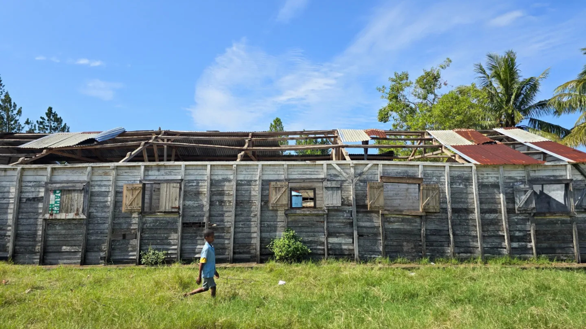 A child walks past a weather-damaged wooden school building with a partially missing roof.