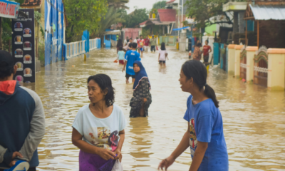 Residents wading through floodwater on a flooded residential street, with homes and buildings on either side.