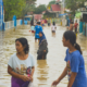 Residents wading through floodwater on a flooded residential street, with homes and buildings on either side.