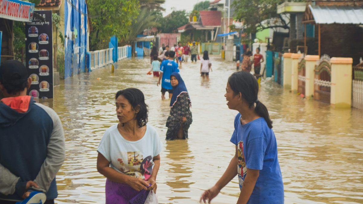 Residents wading through floodwater on a flooded residential street, with homes and buildings on either side.