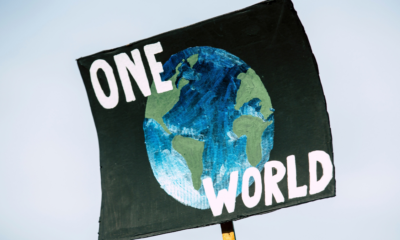 A black climate protest sign reading “ONE WORLD” with a hand-painted illustration of Earth, symbolising global cooperation on climate change ahead of COP31.