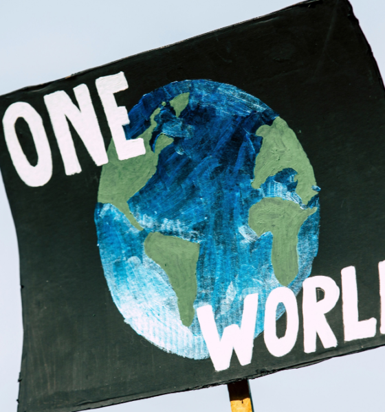 A black climate protest sign reading “ONE WORLD” with a hand-painted illustration of Earth, symbolising global cooperation on climate change ahead of COP31.
