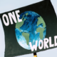 A black climate protest sign reading “ONE WORLD” with a hand-painted illustration of Earth, symbolising global cooperation on climate change ahead of COP31.