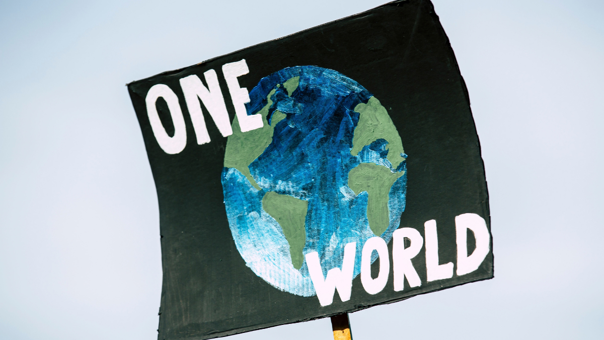 A black climate protest sign reading “ONE WORLD” with a hand-painted illustration of Earth, symbolising global cooperation on climate change ahead of COP31.