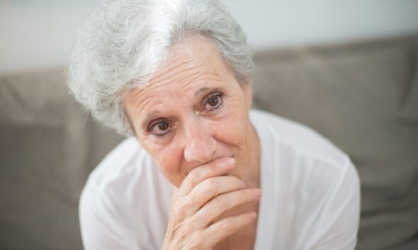 Elderly woman with grey hair sitting indoors with her hand near her mouth and a thoughtful expression.