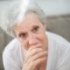 Elderly woman with grey hair sitting indoors with her hand near her mouth and a thoughtful expression.
