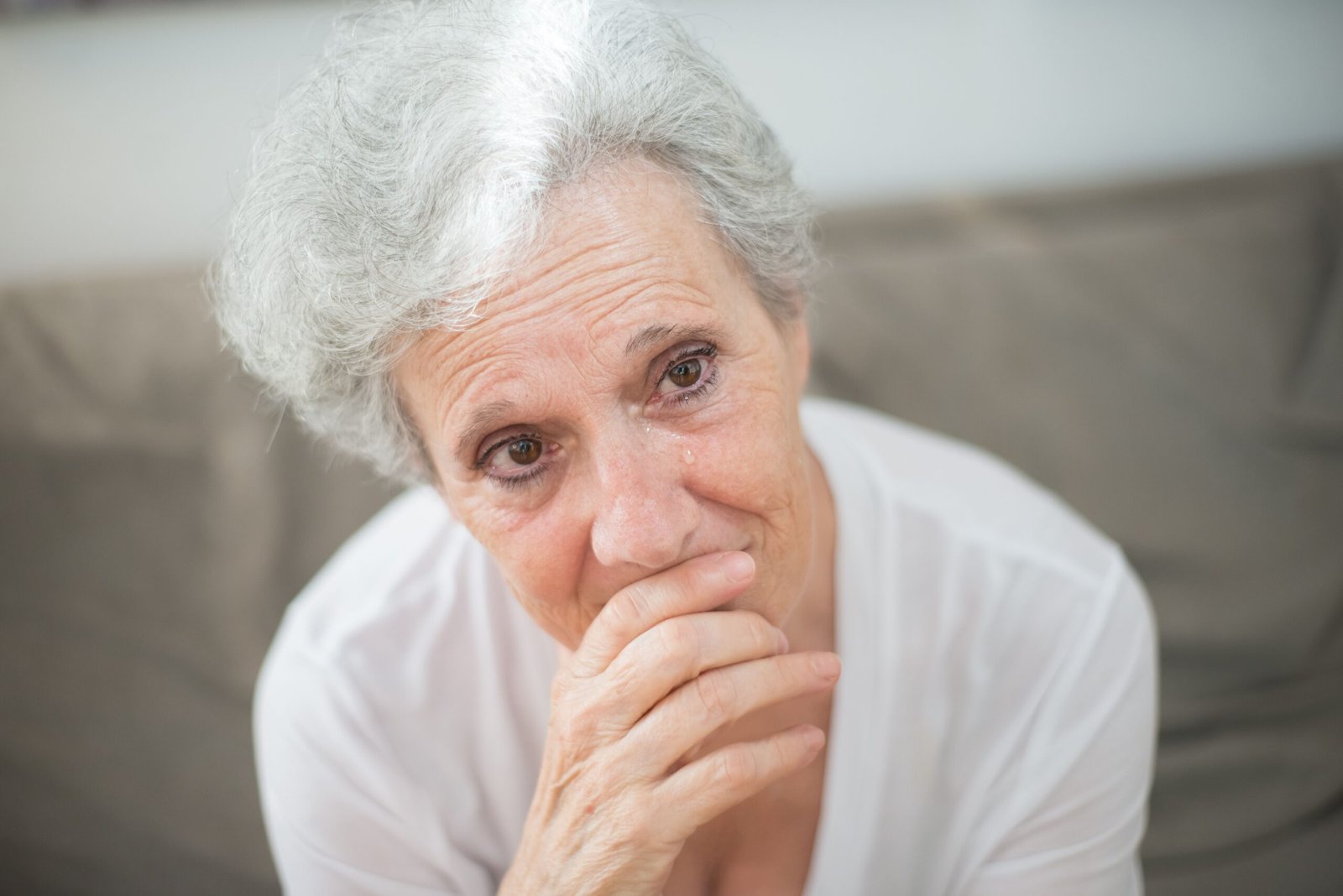 Elderly woman with grey hair sitting indoors with her hand near her mouth and a thoughtful expression.