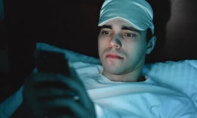 Young man lying in bed wearing a blue sleep mask on his forehead and looking at a smartphone in dim lighting.