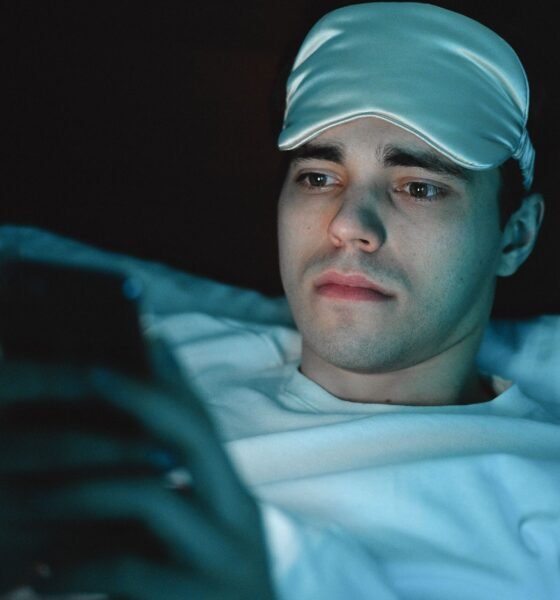 Young man lying in bed wearing a blue sleep mask on his forehead and looking at a smartphone in dim lighting.